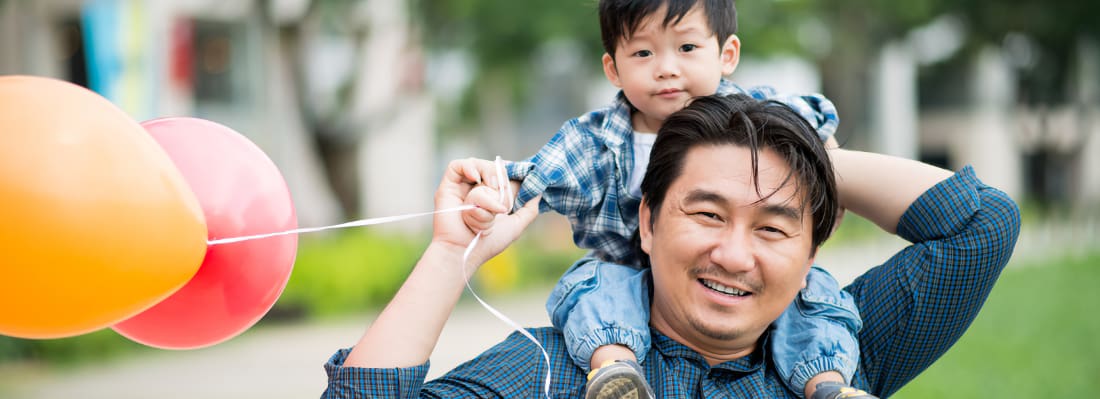 An Asian father hold his toddler on his shoulders, along with a bunch of colorful balloons