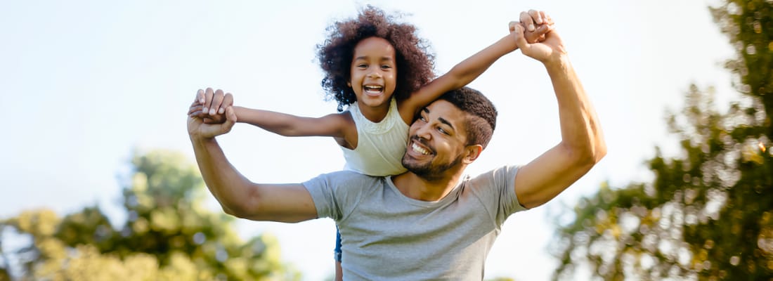 A smiling Black father holds his laughing daughter on his shoulders
