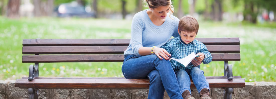A mother sits on a park bench with her young son, helping him read a book